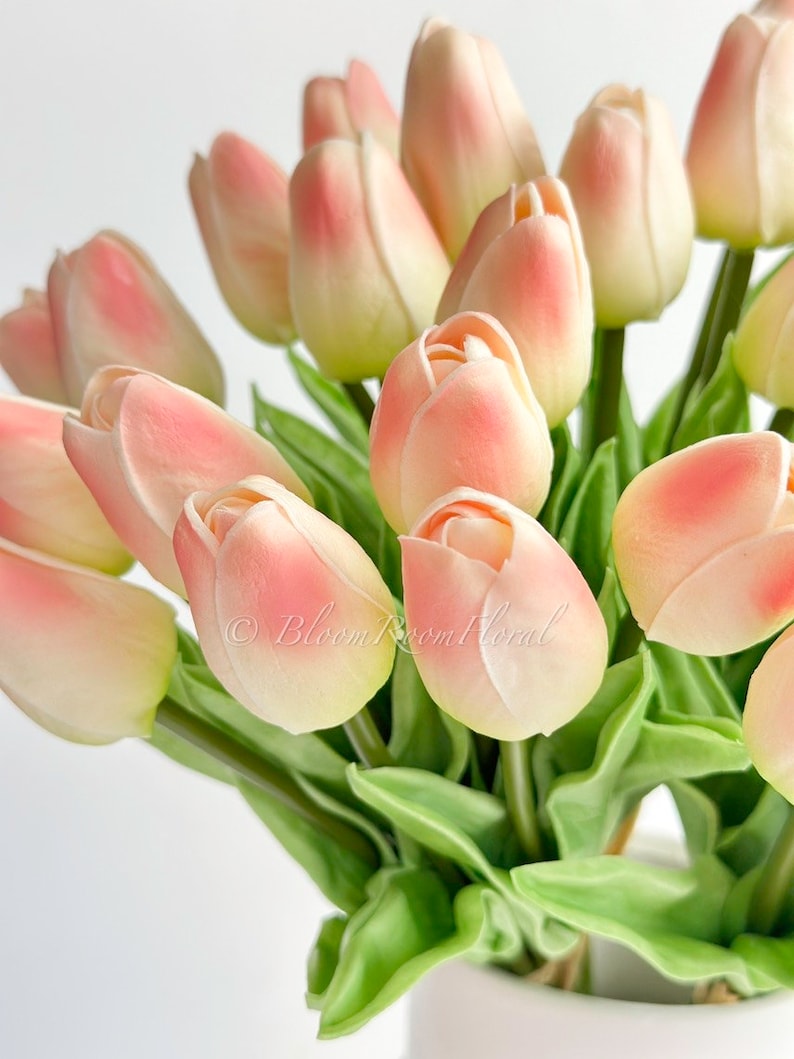 May include: A bouquet of peach-coloured artificial tulips with green leaves in a white vase. The tulips are arranged in a cluster, with some of the flowers facing the camera and others facing away. The leaves are arranged around the tulips, creating a lush and full bouquet.