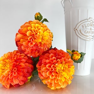 May include: Three orange and yellow dahlia flowers with green stems and leaves. The flowers are in a white vase with the words "Flowers & Garden" on the side.