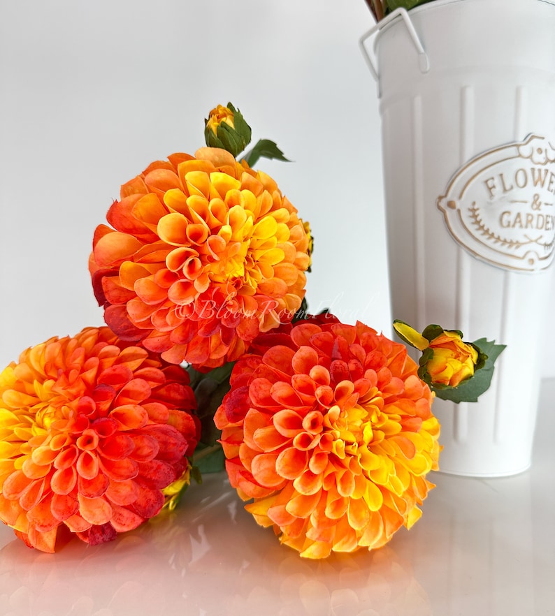 May include: Three orange and yellow dahlia flowers with green stems and leaves. The flowers are in a white vase with the words "Flowers & Garden" on the side.