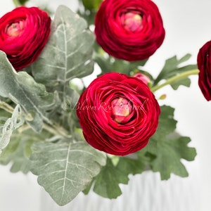 May include: A close-up shot of a bouquet featuring deep red ranunculus flowers, showcasing multiple layers of petals. The flowers are complemented by silvery-green leaves and presented in a white vase. The lighting accentuates the textures and colours of the floral arrangement.