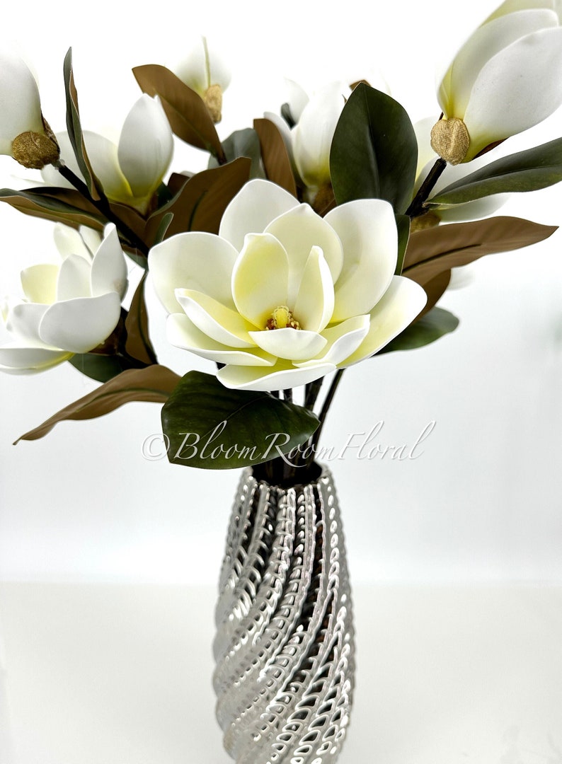 K&ouml;nnte beinhalten: Ein Strau&szlig; k&uuml;nstlicher Magnolienbl&uuml;ten in einer silbernen Vase. Die Bl&uuml;ten sind wei&szlig; mit gelben Zentren, umgeben von dunkelgr&uuml;nen und braunen Bl&auml;ttern. Die Vase hat ein strukturiertes Spiraldesign. Der Text "Bloom Room Floral" ist sichtbar.