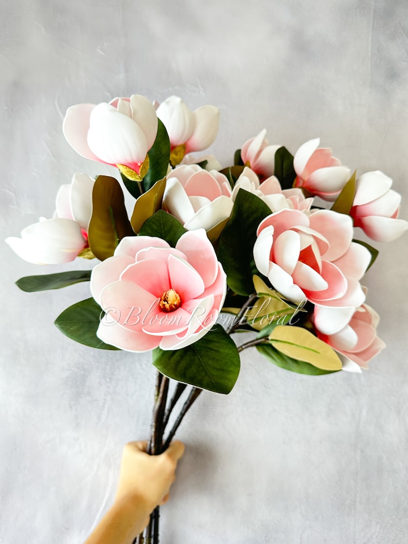 May include: A bouquet of artificial pink and white magnolia flowers with green leaves. The flowers have a delicate, layered appearance and are arranged in a hand-held bunch.