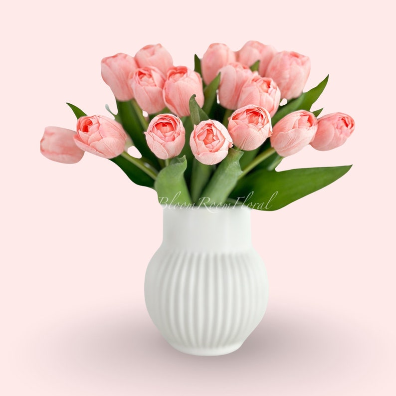May include: A bouquet of pink artificial tulips in a white ceramic vase with vertical ridges. The vase is on a light pink background.
