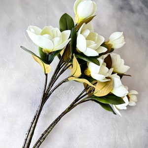 K&ouml;nnte beinhalten: Ein Strau&szlig; k&uuml;nstlicher Magnolienbl&uuml;ten mit gro&szlig;en, cremewei&szlig;en Bl&uuml;tenbl&auml;ttern und dunkelgr&uuml;nen Bl&auml;ttern. Die Stiele sind dunkelbraun. Die Blumen sind in einem nat&uuml;rlichen, eleganten Stil angeordnet, ideal f&uuml;r die Inneneinrichtung.