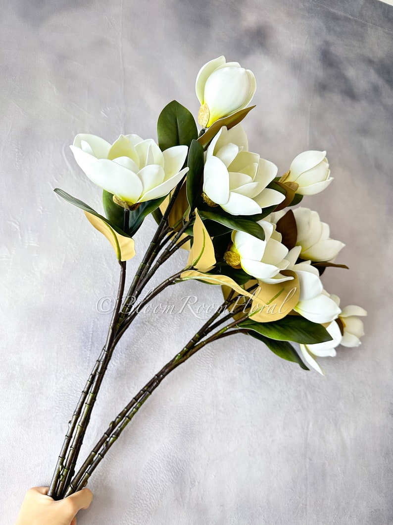 K&ouml;nnte beinhalten: Ein Strau&szlig; k&uuml;nstlicher Magnolienbl&uuml;ten mit gro&szlig;en, cremewei&szlig;en Bl&uuml;tenbl&auml;ttern und dunkelgr&uuml;nen Bl&auml;ttern. Die Stiele sind dunkelbraun. Die Blumen sind in einem nat&uuml;rlichen, eleganten Stil angeordnet, ideal f&uuml;r die Inneneinrichtung.