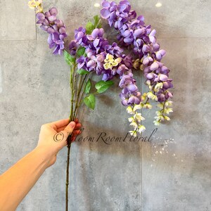 May include: A single stem of artificial wisteria flowers with purple blooms and green leaves. The flowers are arranged in a cascading style, with the stem extending downwards. The flowers are against a gray tiled background.
