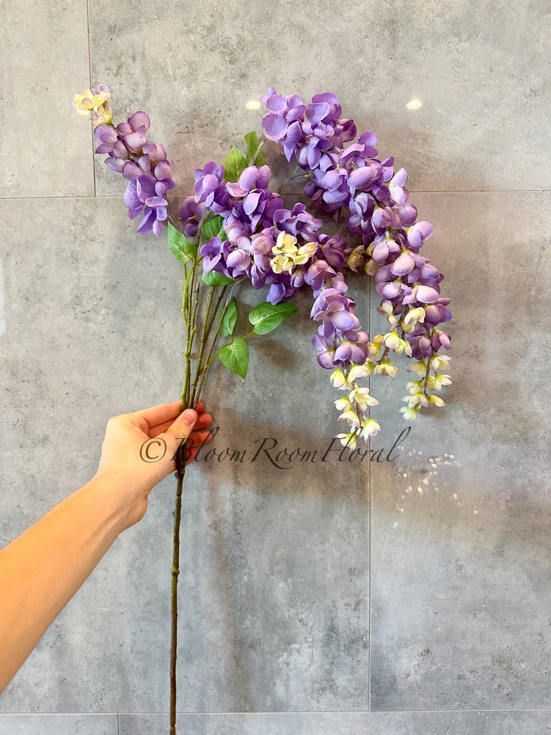 May include: A single stem of artificial wisteria flowers with purple blooms and green leaves. The flowers are arranged in a cascading style, with the stem extending downwards. The flowers are against a gray tiled background.