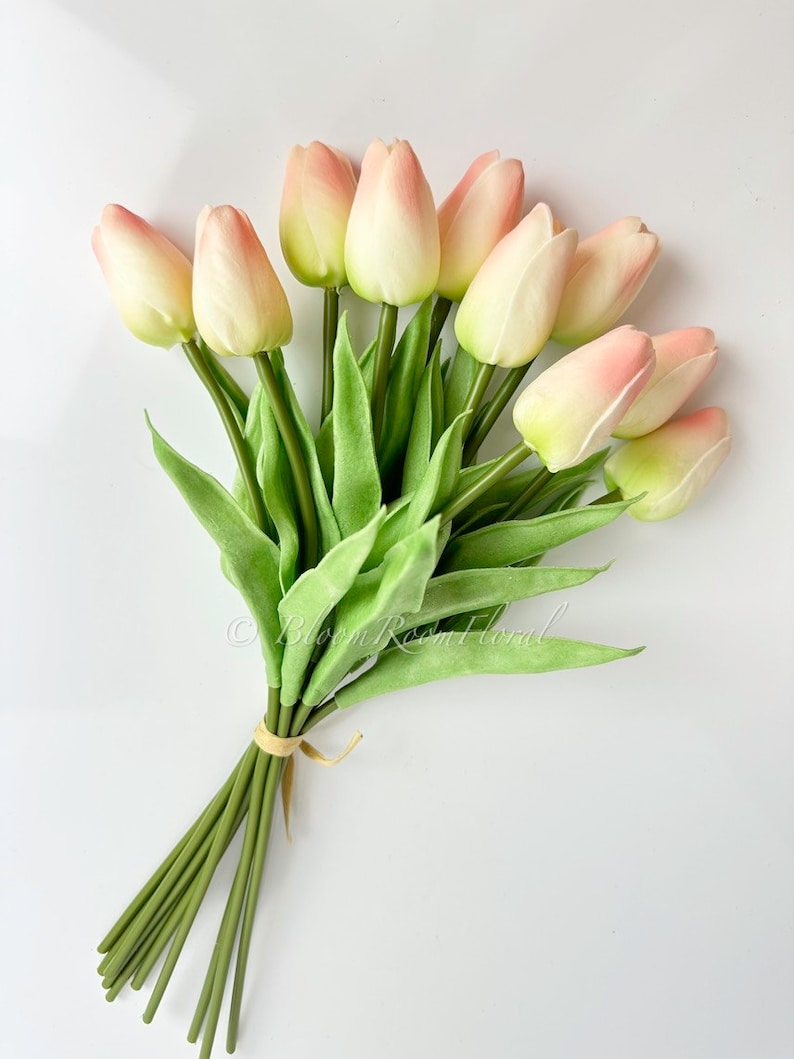 May include: A bouquet of 10 faux pink and white tulips with green stems and leaves. The stems are tied together with a beige ribbon.