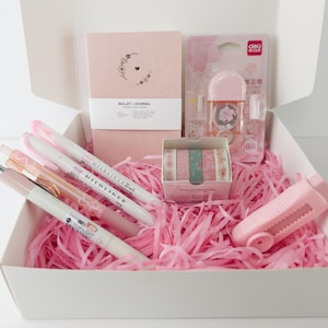 May include: A pink and white gift box filled with stationery supplies, including a pink notebook with the words "BULLET JOURNAL" on the cover, pink pens, washi tape, a pink tape dispenser, and pink tissue paper.