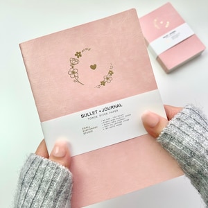 Peut inclure: Un carnet de notes rose avec un motif floral doré et un cœur. Le carnet est fabriqué avec du papier Tomoe River et a une couverture en cuir PU souple. Il contient 180 pages blanches.