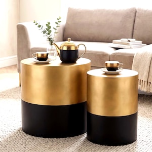 May include: Two cylindrical side tables, one large and one small, with a gold upper half and a black lower half. A black and gold teapot and teacups sit on top. The tables are in front of a beige sofa.