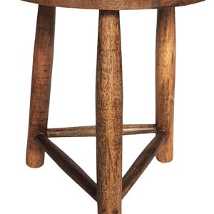 May include: A small, round wooden stool with three legs. The stool has a natural wood finish with a dark, decorative design on the top surface. The legs are connected by a lower support structure.