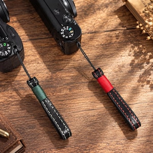 May include: Two black leather camera wrist straps attached to black digital cameras. One strap features a dark green fabric accent with white stitching, while the other strap has a bright red fabric accent with red stitching along the leather edges.