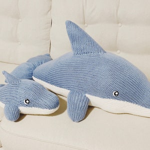 May include: Two knitted blue dolphin plush toys. The larger dolphin is lying down with its head turned to the side. The smaller dolphin is lying on its side with its head facing forward. Both dolphins have white bellies and black eyes.