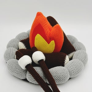 Op de afbeelding: Een gebreid pluche kampvuur met een rode en oranje vlam, twee bruine boomstammen en twee witte marshmallows.