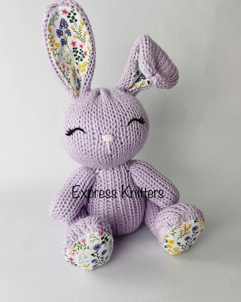 May include: A lavender knitted bunny with floral fabric accents on the ears and feet. The bunny has a sweet expression with embroidered eyes and a pink nose.