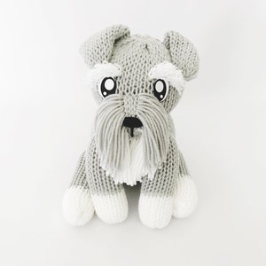 Könnte beinhalten: Ein graues, gestricktes Plüschtier in Form eines Hundes mit weißen Felldetails, großen schwarzen Augen und einer schwarzen Nase. Der Hund sitzt auf einem weißen Hintergrund.