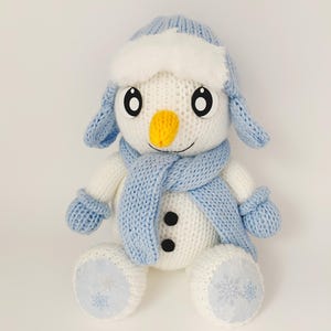 Könnte beinhalten: Ein handgefertigtes Schneemann-Plüschtier mit weißem Körper, blauer Mütze, Schal und Fäustlingen. Der Schneemann hat eine gelbe Nase, schwarze Knopfaugen und einen lächelnden Mund. Die Füße haben ein Schneeflocken-Design.