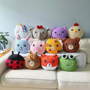 May include: A collection of eleven handmade, round animal-shaped pillows. The pillows include a unicorn, elephant, cat, chicken, bunny, pig, giraffe, bear, ladybug, llama, fox, and frog. Each pillow features detailed facial features and is displayed on a white couch.