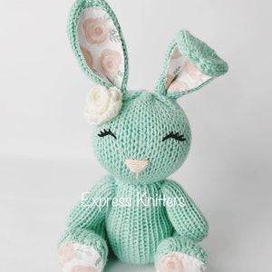 May include: A hand-knitted green bunny with floral fabric ears and feet. The bunny has a white flower on its head and embroidered eyes and nose. The bunny is sitting on a white background. Express Knitters is written on the bunny's belly.