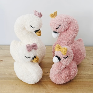Könnte beinhalten: Drei gestrickte Plüschtiere in Form von Schwänen. Zwei sind weiß mit rosa Schleifen und eines ist rosa mit einer gelben Schleife. Alle drei haben goldene Kronen auf dem Kopf.