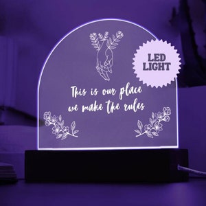 May include: A white LED light with a wooden base. The light features a design of two hands holding each other with flowers and leaves. The text "This is our place we make the rules" is written below the hands. The light is illuminated in a purple hue.