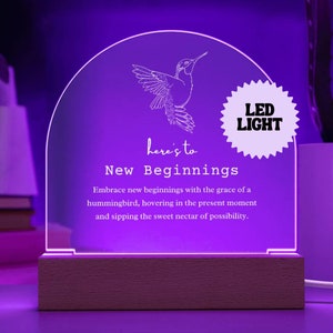 Może przedstawiać: Fioletowe światło LED z drewnianą podstawą przedstawia wzór kolibra i tekst "here's to New Beginnings". Tekst kontynuuje: "Embrace new beginnings with the grace of a hummingbird, hovering in the present moment and sipping the sweet nectar of possibility."