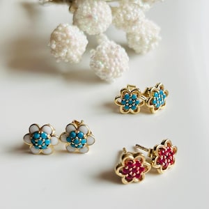 Op de afbeelding: Drie paar gouden oorbellen met een bloemvorm. Een paar heeft wit emaille met turquoise stenen, een paar heeft blauw emaille met turquoise stenen en een paar heeft rood emaille met rode stenen.
