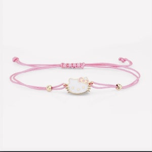 Può includere: Un braccialetto di corda rosa con un ciondolo Hello Kitty dorato. Il ciondolo è bianco con dettagli rosa e un fiocco dorato.