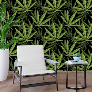May include: A white chair with black metal legs sits in front of a black wall with a green marijuana leaf pattern. A small black side table with a book and a potted plant sits next to the chair.