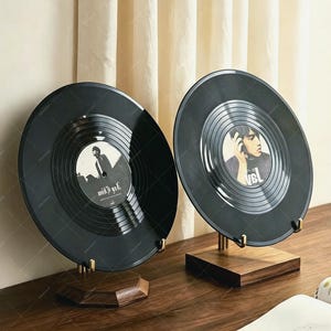May include: Two black vinyl records displayed on individual wooden stands. Each record features a central circular label with a black and white photographic portrait of a person. The records are positioned upright, showcasing the glossy grooved surfaces.