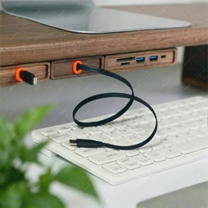 May include: A wooden monitor stand featuring an integrated USB hub with multiple ports, including SD card slots and USB-A inputs. The front panel displays two bright orange circular accents around cable ports, with a black charging cable plugged in.
