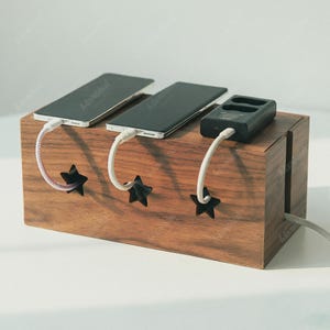 Può includere: Una stazione di ricarica in legno con tre smartphone e un piccolo dispositivo nero sopra. La stazione ha ritagli a forma di stella per i cavi di ricarica. Il legno ha una calda tonalità marrone.