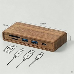 Puede incluir: Un hub USB rectangular de madera con dos puertos USB-A, un puerto USB-C y dos ranuras para tarjetas de memoria. El dispositivo mide 112 mm por 48 mm por 20 mm. Debajo del hub hay iconos de l&iacute;neas que representan una tarjeta SD y tres cables.