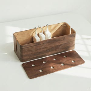 May include: A wooden cable organizer with a rectangular shape and a removable lid. The organizer is made of dark brown wood and has several holes for cables. Three white charging adapters are visible inside the organizer.