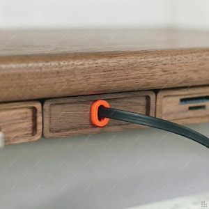 May include: A dark brown wooden desk edge featuring a rectangular panel with a bright orange circular grommet. A thin black cable passes through the center of the orange grommet, extending outward from the wooden surface of the desk structure.