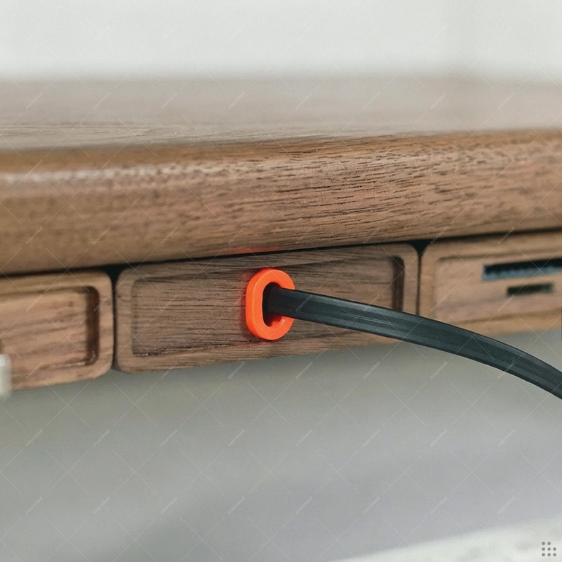 May include: A dark brown wooden desk edge featuring a rectangular panel with a bright orange circular grommet. A thin black cable passes through the center of the orange grommet, extending outward from the wooden surface of the desk structure.