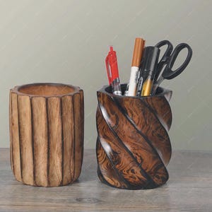 Puede incluir: Dos organizadores de escritorio de madera. Uno es un portalápices cilíndrico con estrías verticales. El otro es un portalápices tallado y retorcido con bolígrafos, rotuladores y tijeras en su interior. Ambos son de madera con acabado natural.
