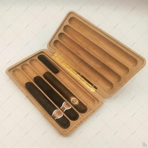 Puede incluir: Estuche de puros de madera abierto con cuatro puros visibles. El estuche es de color marrón claro con una bisagra dorada. Los puros son de color marrón oscuro con etiquetas blancas y doradas. También hay un cortapuros negro.