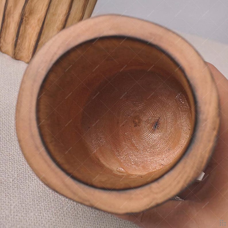 May include: A wooden mortar and pestle set, viewed from above. The mortar is a cylindrical bowl with a dark brown interior and a lighter brown exterior. The wood grain is visible. The pestle is not visible.