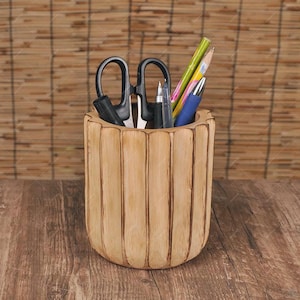 May include: A light brown wooden desk organizer with vertical grooves, holding black scissors, pens, and pencils. The organizer sits on a wooden surface, with a bamboo blind in the background.