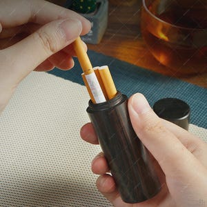 May include: A dark brown wooden cigarette case with a cylindrical shape, holding several cigarettes. The case has a lid and is being held by a person. The cigarettes are yellow and white.