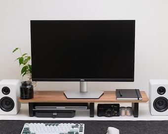 May include: A white desk with a black monitor, a white keyboard, a white mouse, and two white speakers. There is a wooden monitor stand with a black notebook, a camera, and other items on it. The desk has a black mouse pad and a green plant in a pot on the left side.