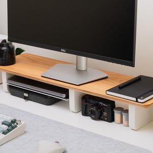 May include: A monitor stand featuring a light-toned wood grain top surface supported by two white vertical legs. The stand provides storage space underneath for a laptop, a black camera, and a notebook, while supporting a black Dell computer monitor.