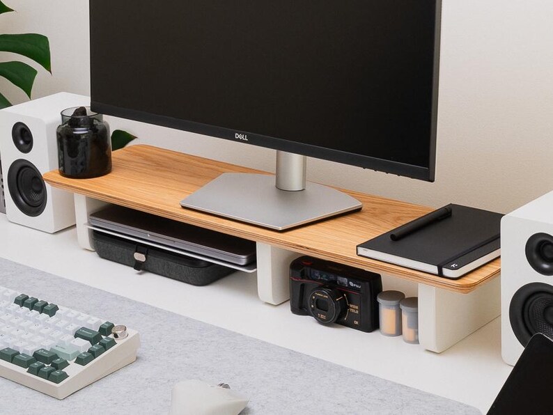 May include: A monitor stand featuring a light-toned wood grain top surface supported by two white vertical legs. The stand provides storage space underneath for a laptop, a black camera, and a notebook, while supporting a black Dell computer monitor.