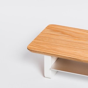 May include: A rectangular monitor stand featuring a light brown wood grain top surface with rounded corners. The stand is supported by two solid white vertical legs and includes a thin, flat, metallic silver shelf positioned between the legs.
