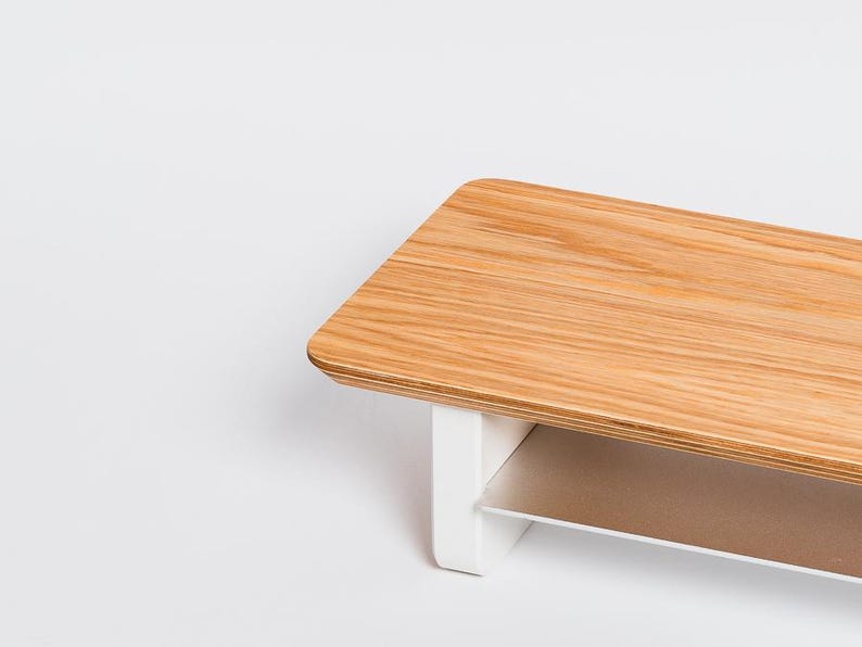 May include: A rectangular monitor stand featuring a light brown wood grain top surface with rounded corners. The stand is supported by two solid white vertical legs and includes a thin, flat, metallic silver shelf positioned between the legs.