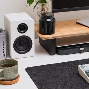 May include: A gray CalDigit docking station, a white speaker with black drivers, a green ceramic mug on a cork coaster, a black glass jar, a wooden monitor stand, and a mechanical keyboard with green and white keys on a dark felt desk mat.