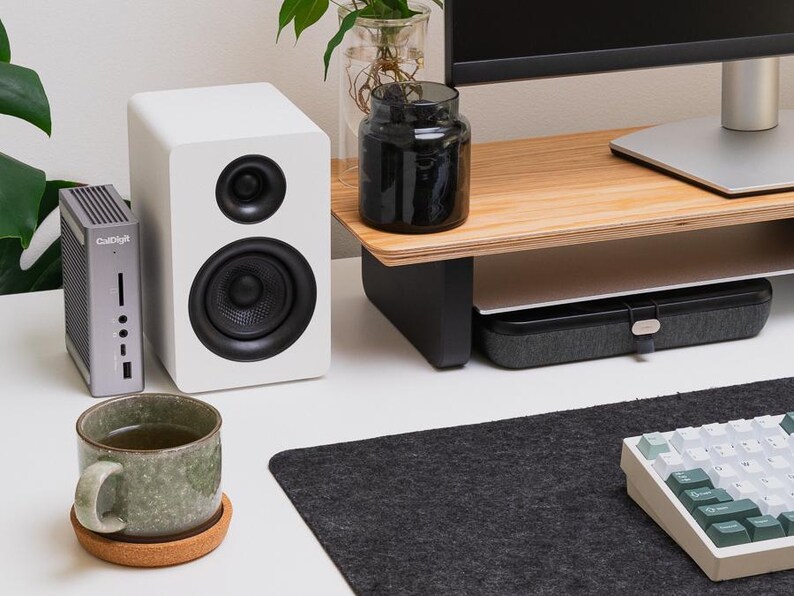 May include: A gray CalDigit docking station, a white speaker with black drivers, a green ceramic mug on a cork coaster, a black glass jar, a wooden monitor stand, and a mechanical keyboard with green and white keys on a dark felt desk mat.