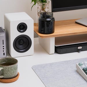 May include: A gray CalDigit docking station, a white speaker with a black driver, a green speckled ceramic mug on a round cork coaster, and a mechanical keyboard with white and green keys arranged on a desk with a monitor stand.